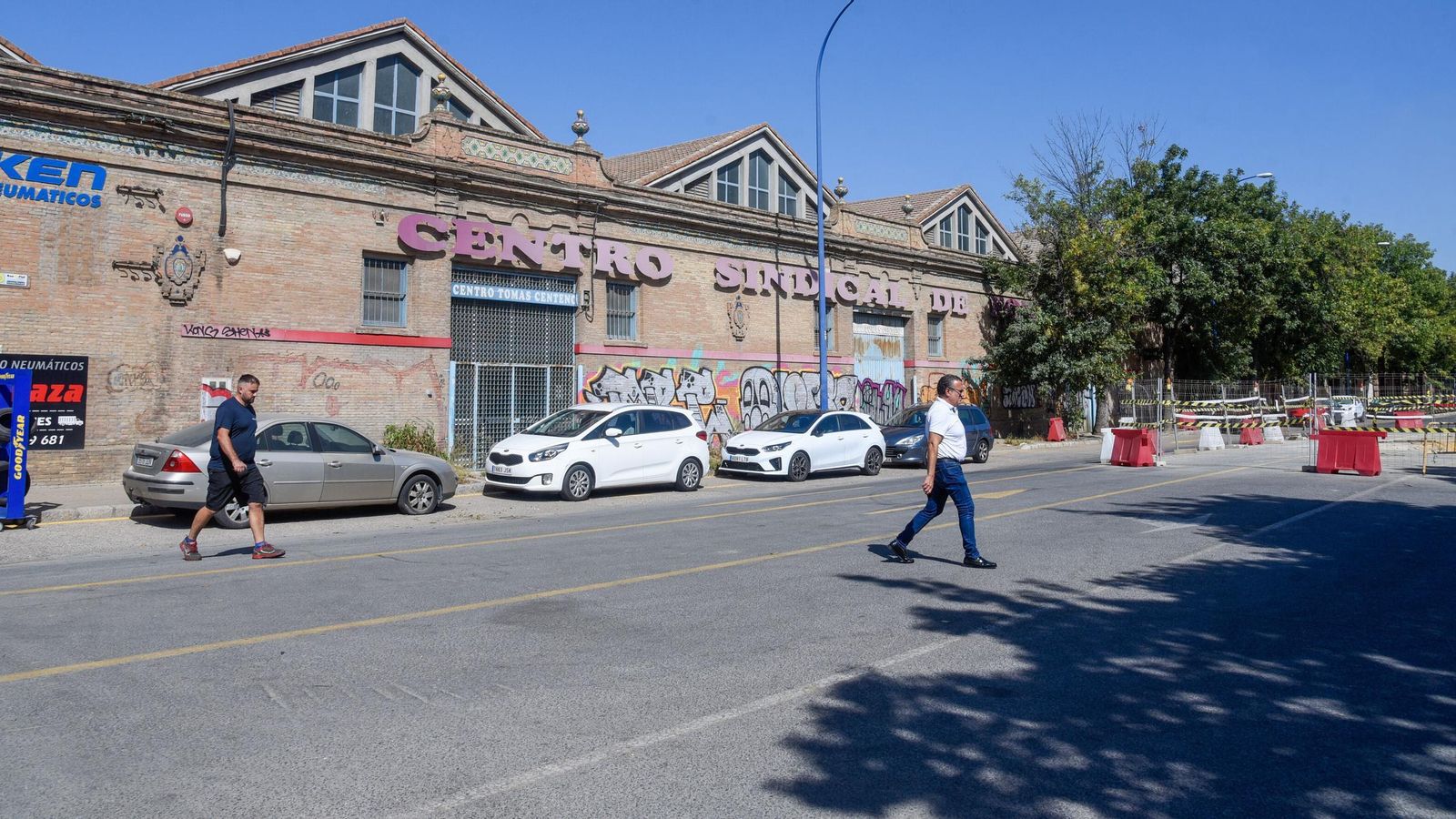 Dos personas cruzan la calzada con las naves de la avenida de las Razas al fondo.