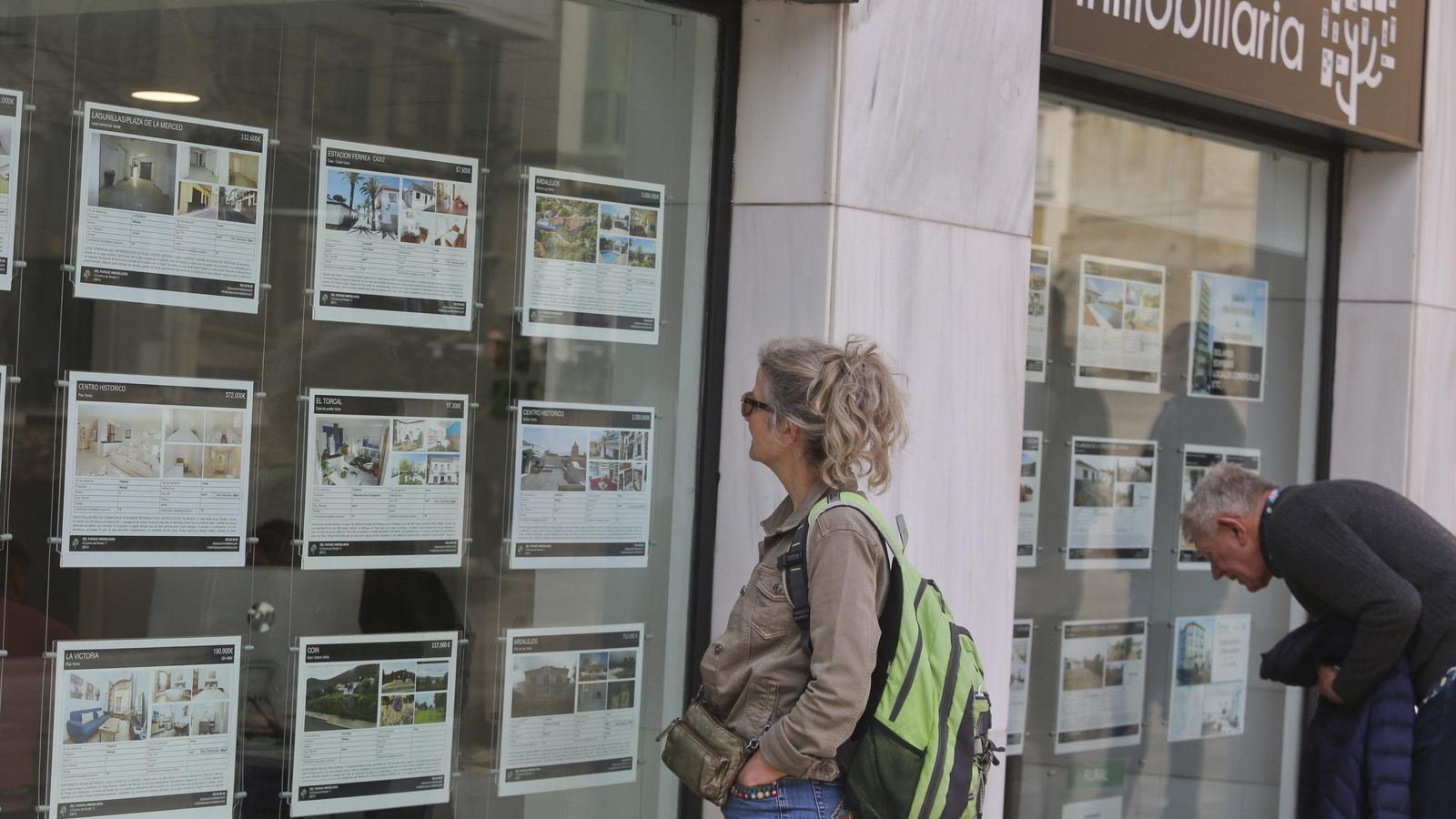 Dos personas miran precios en el escaparate de una inmobiliaria.
