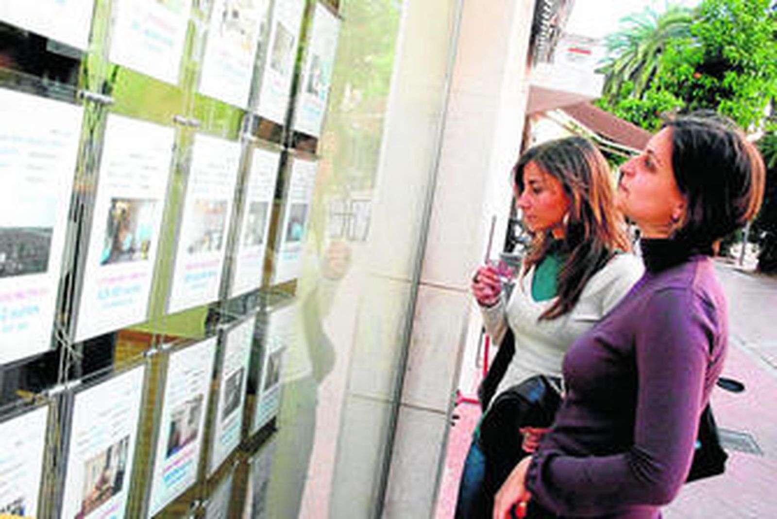 Dos jóvenes miran los anuncios de viviendas en venta en el escaparate de una inmobiliaria de la capital onubense.