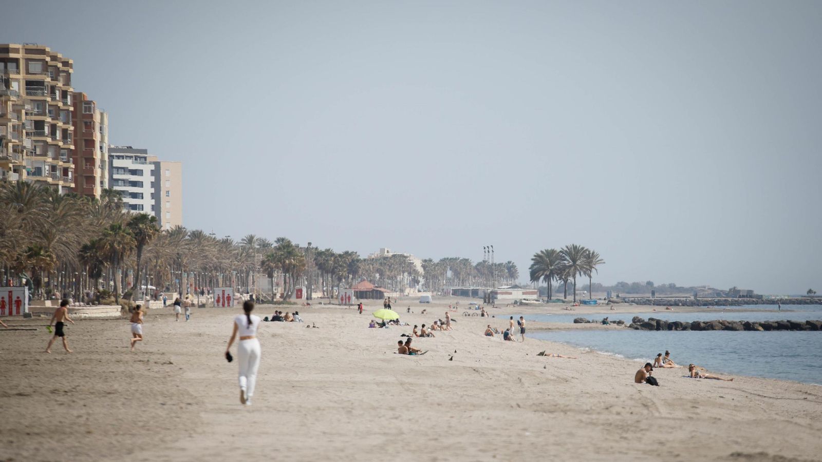 Una chica pasea por una playa de Almería mientras otras personas toman sol tumbadas en la arena.