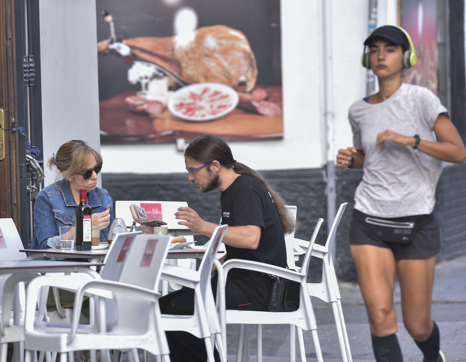 Una joven hace deporte cerca de una pareja sentada en una terraza de la capital hispalense.