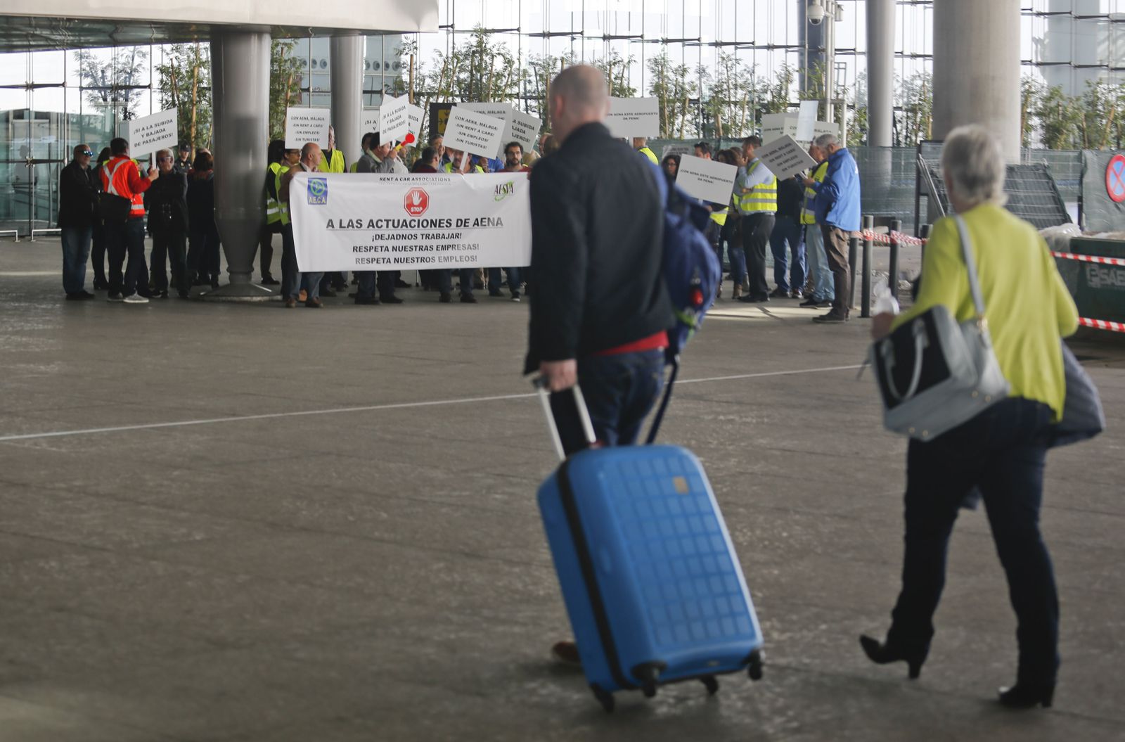 Dos turistas entran ayer en el aeropuerto con la concentración de los empresarios al fondo.