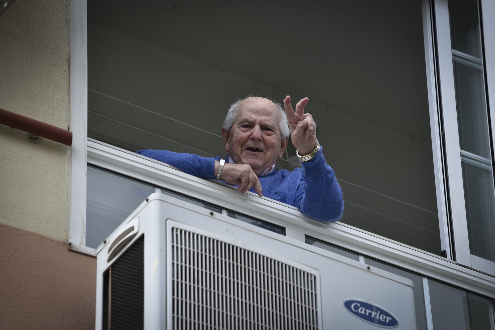 Un hombre mayor muestra el signo de la victoria desde el balcón de su casa.