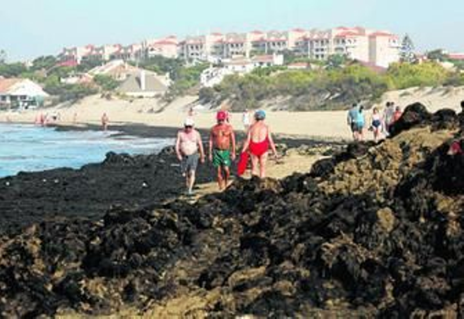 Un grupo de bañistas pasa junto a una montonera de algas, entre la playa de La Bota y El Portil.