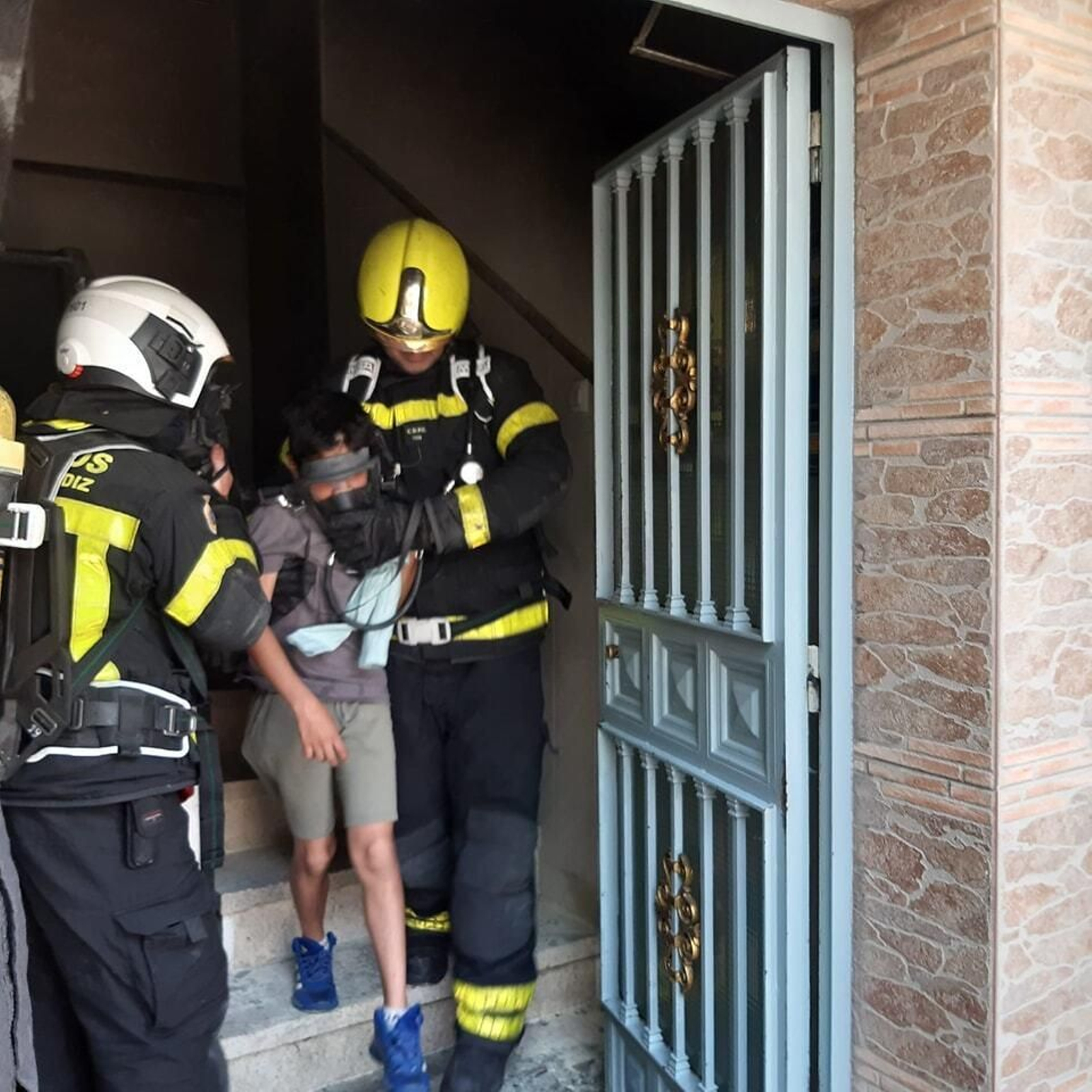 Uno de los menores abandona el bloque con mascarilla y acompañado por bomberos del Parque de Jerez.