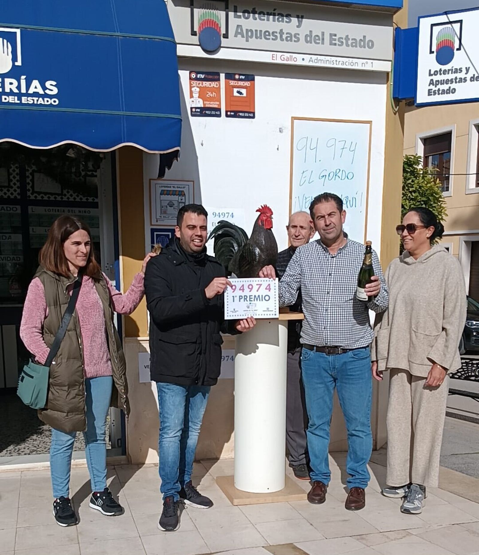 Julián Torralba (con la botella de champán) posa junto a su despacho de lotería, El Gallo, en Morón.