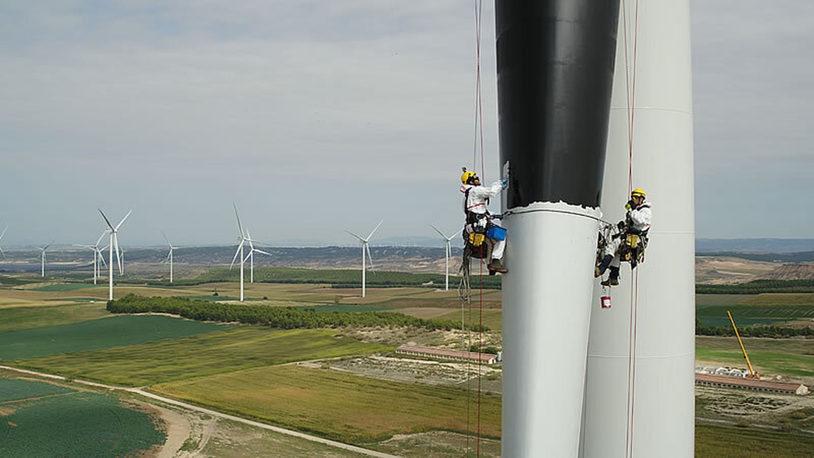 Dos operarios pintan de negro las palas de un aerogenerador.