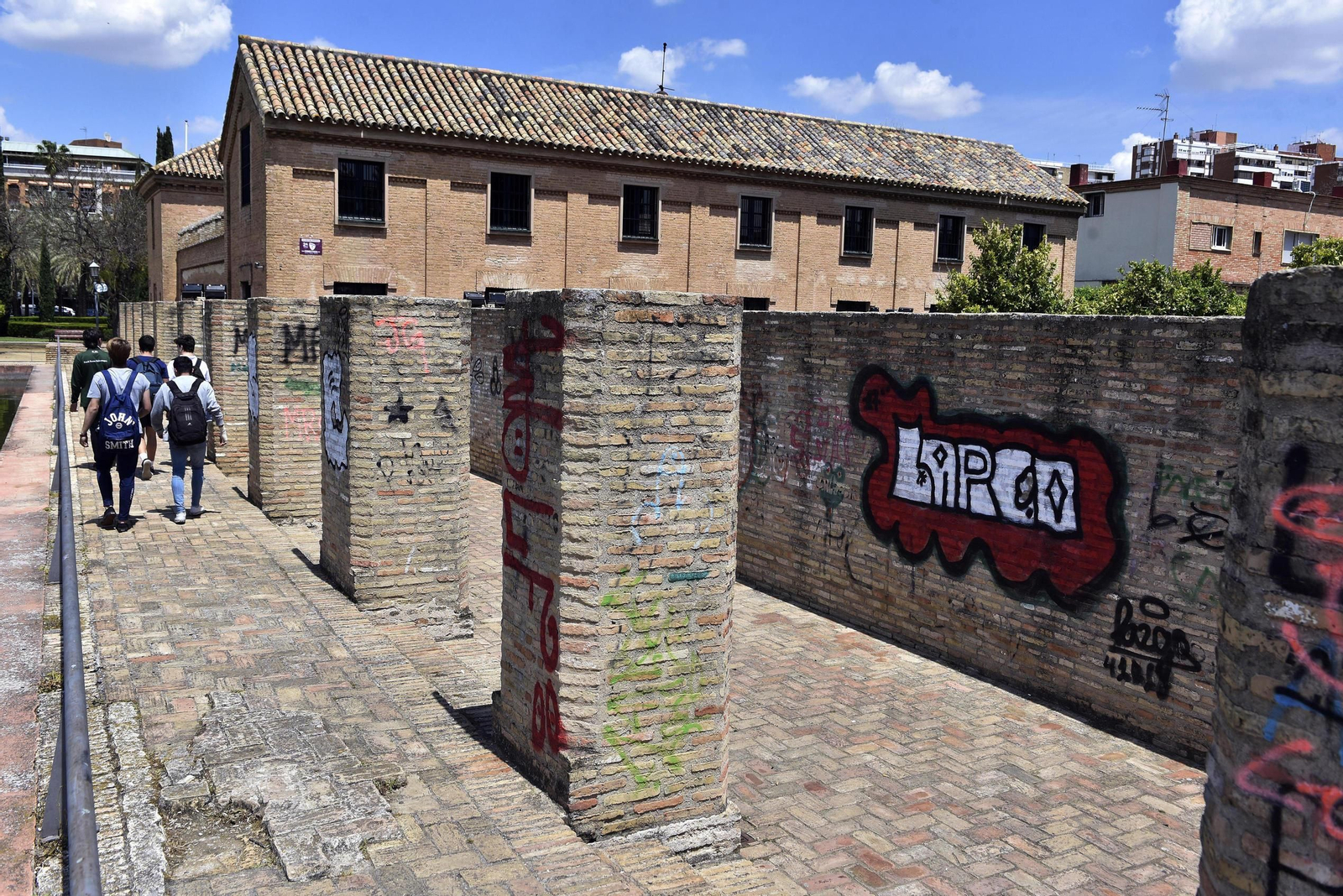 1. Los restos del antiguo palacio almohade completamente cubierto por pintadas. 2. Grafitis en una de las pérgolas del extremo oeste del jardín. 3. Pasarela para recorrer el palacio en mal estado. 4. Más pintadas en otra zona. 5. Uno de los bancos de madera en mal estado. 6. Dos turistas cruzan el parque. 7. Una alberca llena de basura y restos de ramas caídas.