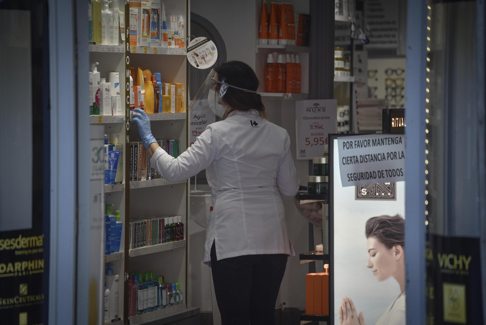 Una farmacéutica provista de mascarilla y pantalla protectora coloca productos en una estantería.