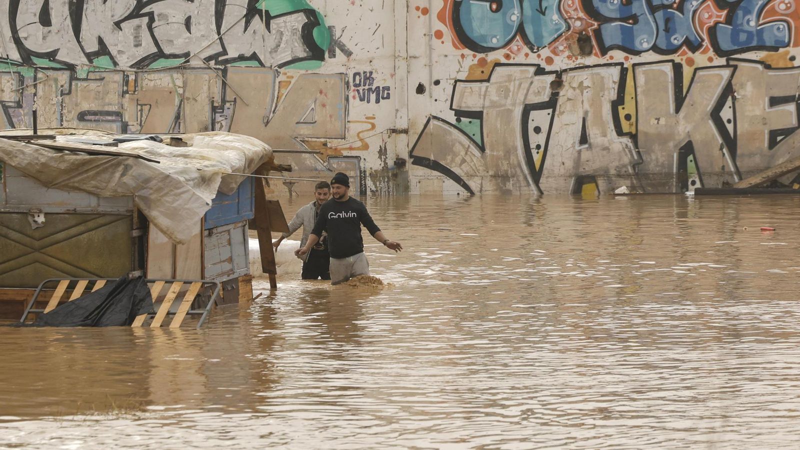 os personas vadean un terreno de chabolas inundado junto a la V-30 en Valencia