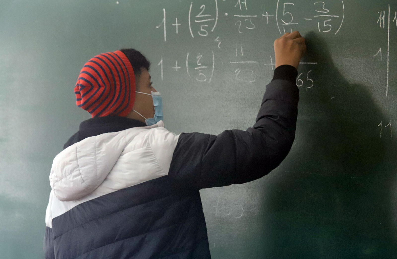 Un alumno realiza una operación en la pizarra con chaquetón y gorro para mitigar el frío que entra por la ventana abierta para la ventilación del aula.