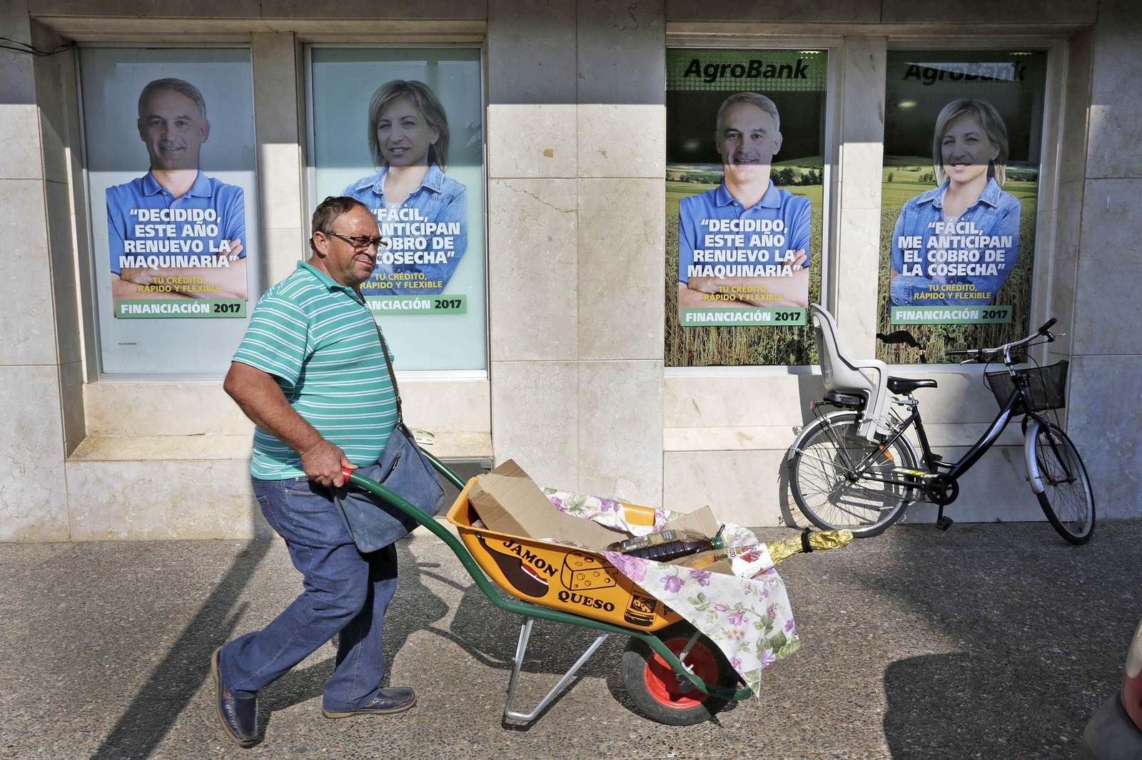 Un vecino de La Barca de la Florida, con un carrillo de mano donde porta los productos de una rifa, pasa frente a la gran sucursal de La Caixa en esta pedanía jerezana.
