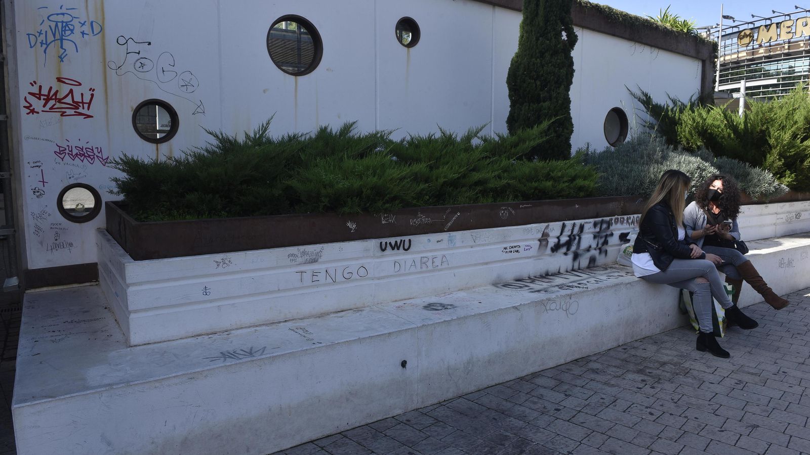 Dos mujeres sentadas en un banco lleno de grafitis al igual que la pared.