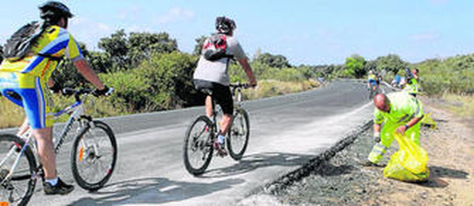 Dos ciclistas circulan por el kilómetro 28 de la A-8002, donde se produjo el accidente, mientras un operario limpia los restos del siniestro.