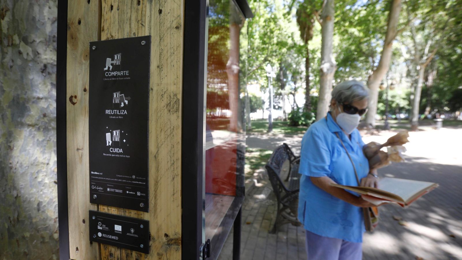 Una señora consulta un libro de la estantería ubicada en los Jardines de Colón.