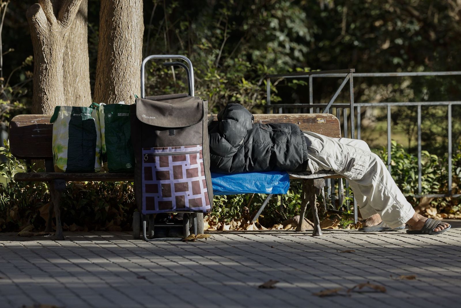 Una persona sin hogar descansa junto a sus pertenencias en un banco público.