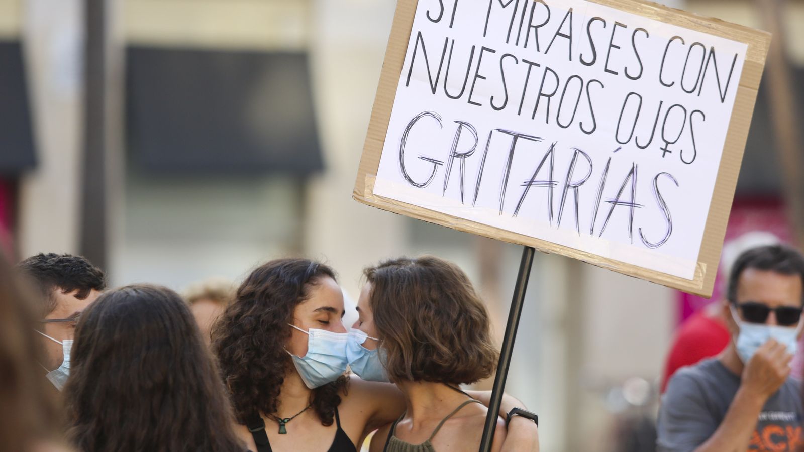 Dos chicas se besan a través de la mascarilla mientras una de ellas sujeta una pancarta.