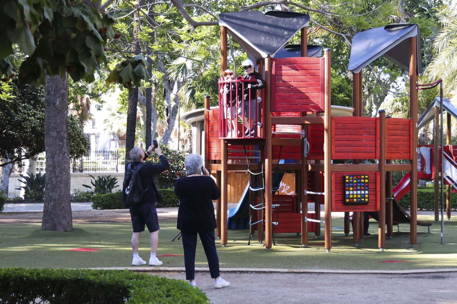 Dos personas mayores fotografían a dos pequeños en un parque infantil.