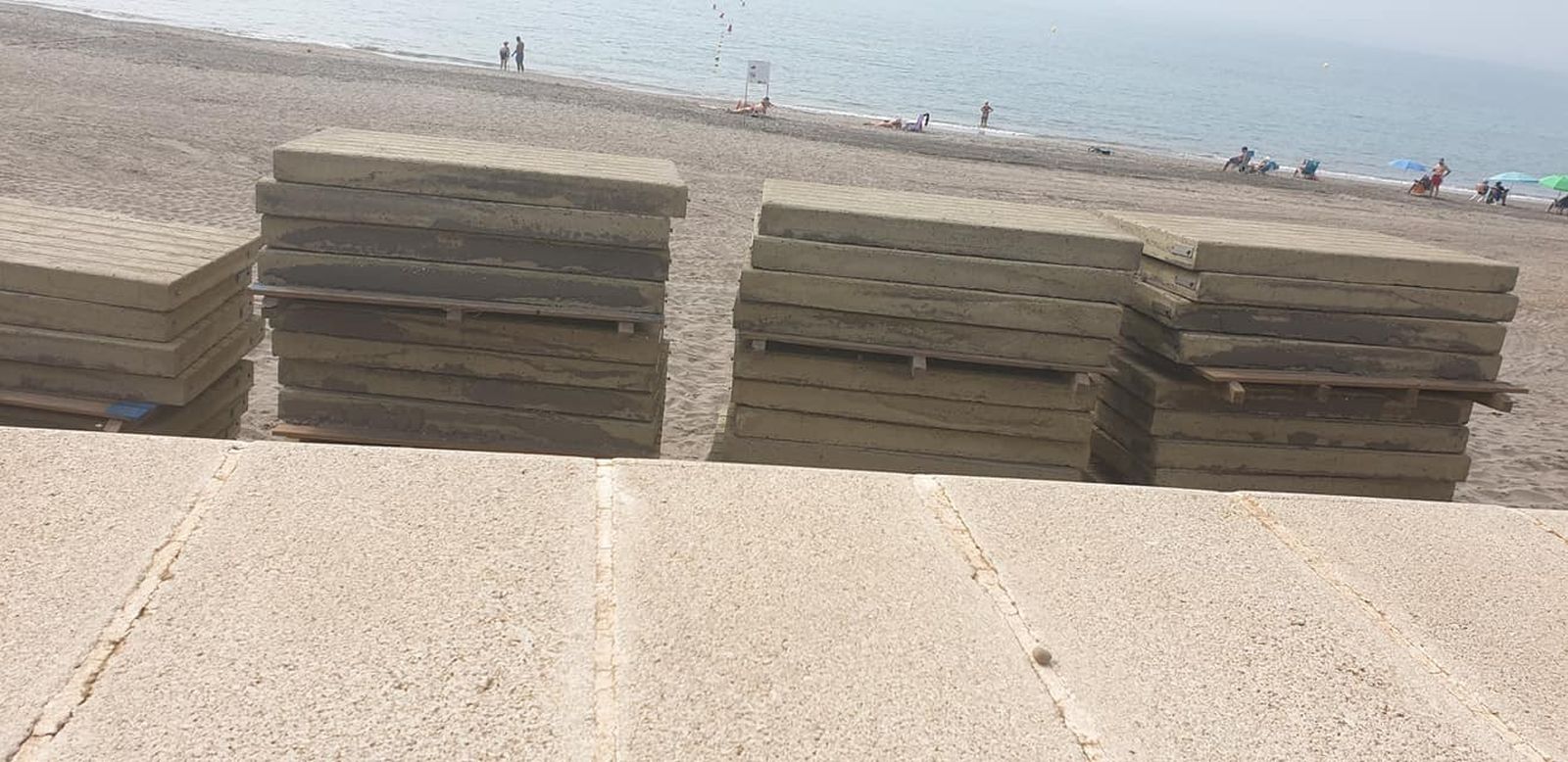 Pasarelas de hormigón desmontadas y apiladas en el paseo marítimo. Al fondo, los bañistas siguen disfrutando de la excelente temperatura.