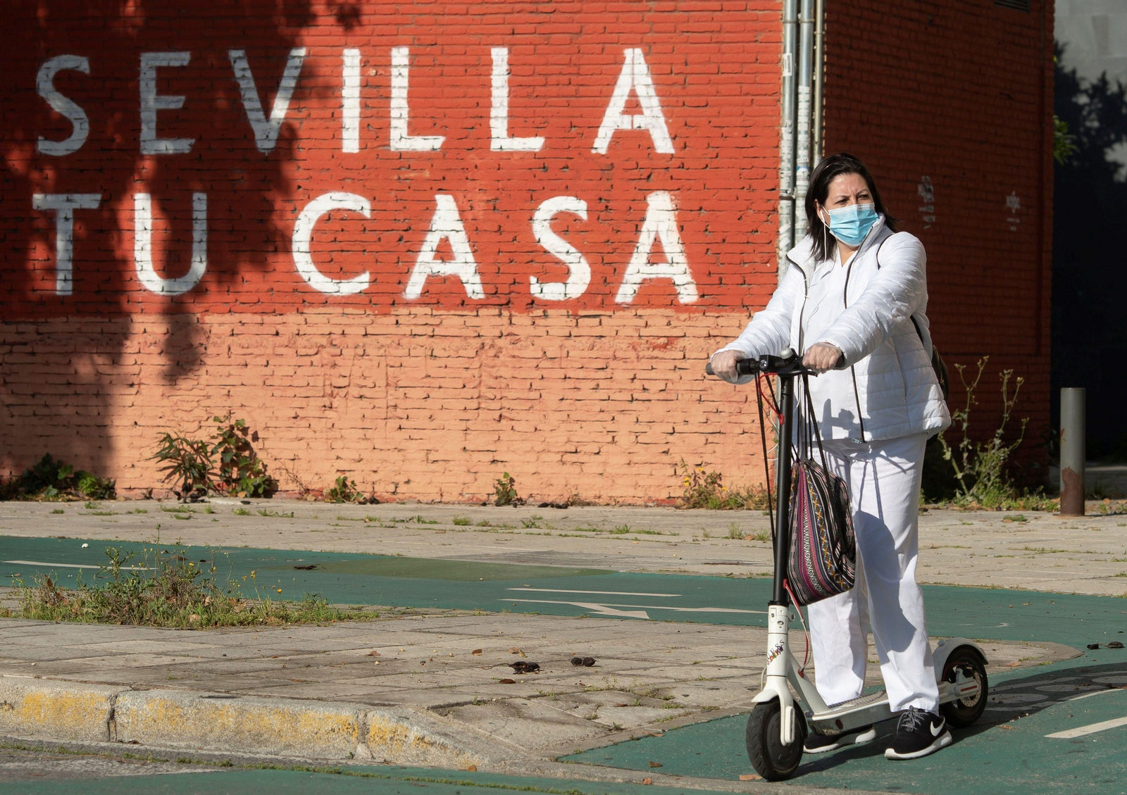 Una mujer protegida con mascarilla y sobre un patinete ante un cartel de 'Sevilla Tu Casa'