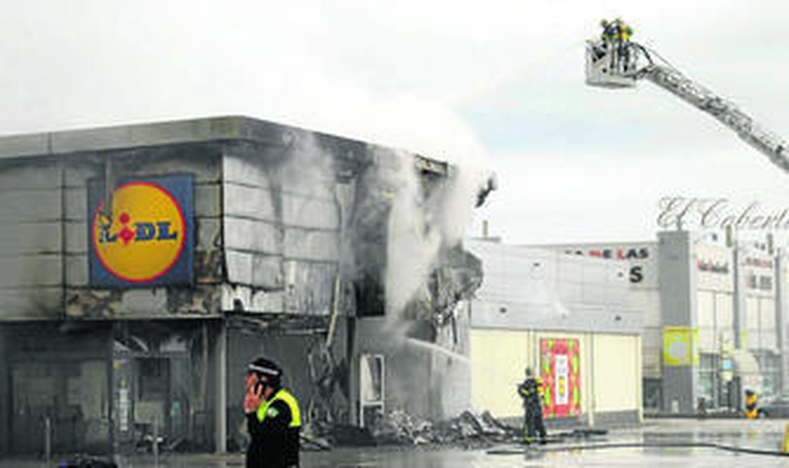 Dos bomberos rocían con agua la tienda afectada por las llamas.