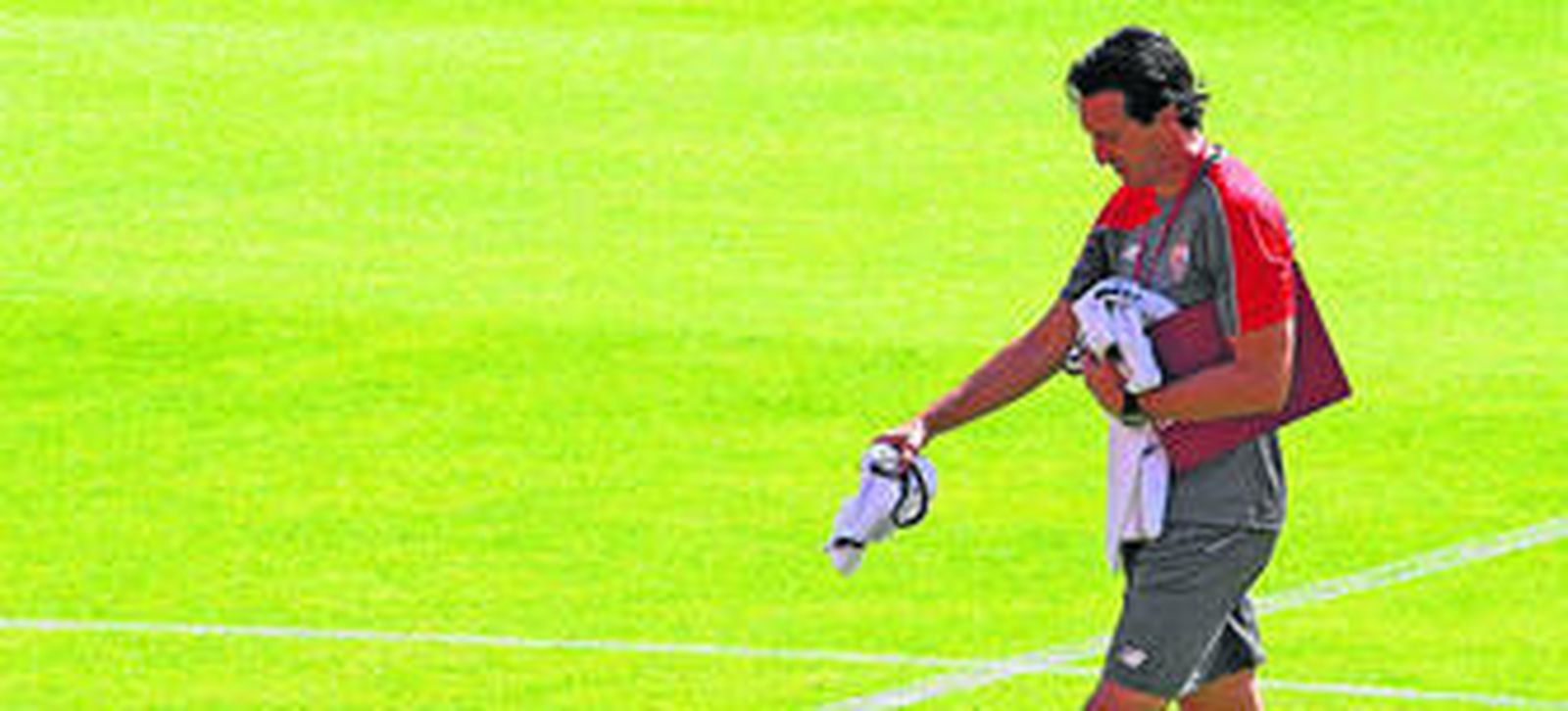 Unai Emery, concentrado, camina sobre el césped portando su carpeta y varios petos para la organización de una tarea de entrenamiento.