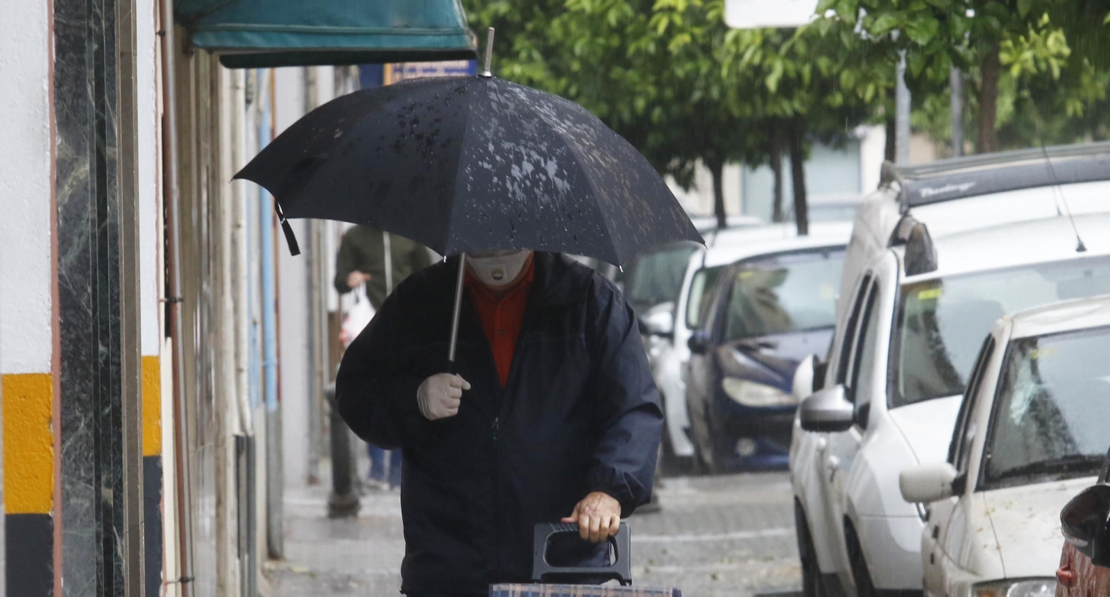 Un hombre se refugia de la lluvia bajo su paraguas a la vez que lleva mascarilla y guantes.