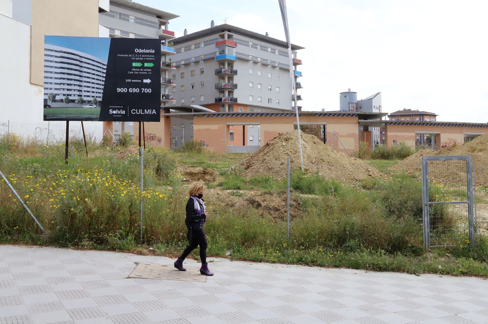 Una mujer pasa delante del solar en el que se construirá la promoción de viviendas que ya anuncia un gran cartel frente a la plaza Ivonne Cazenave.