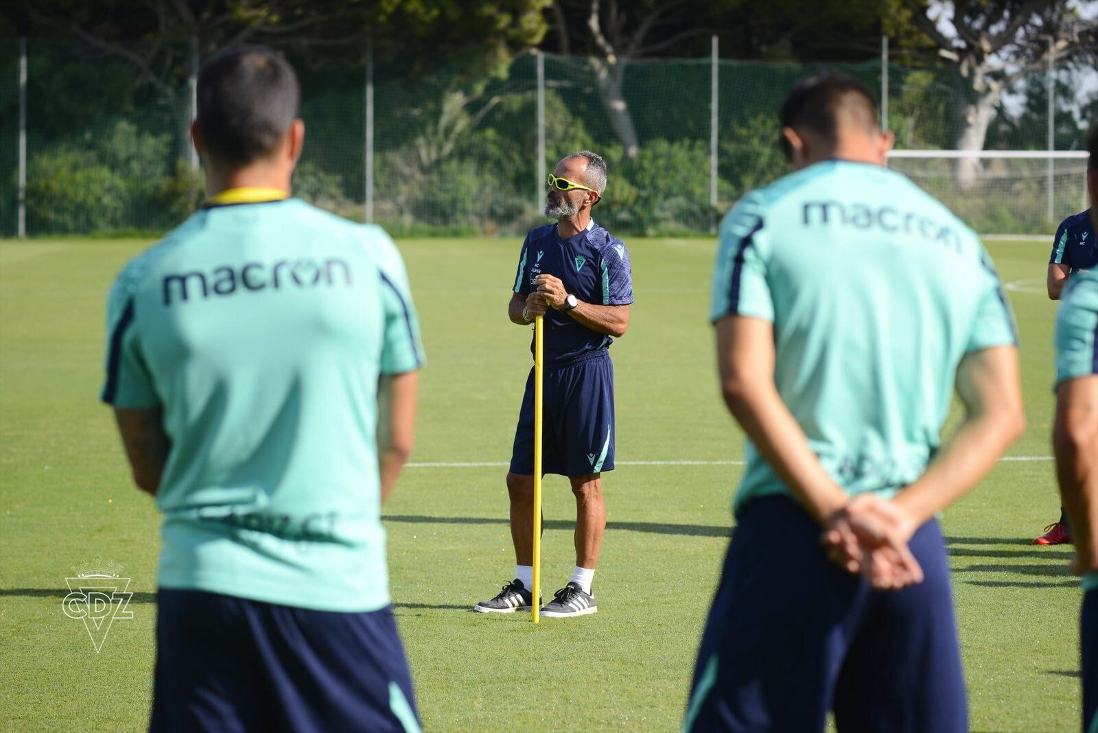 El entrenador se apoya en una pica mientras habla con sus jugadores.