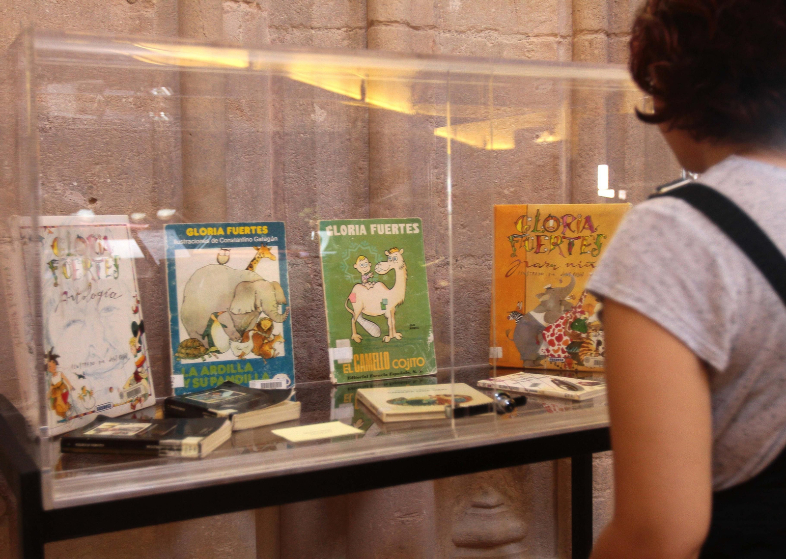 1. María Isabel Montoya sostiene el catálogo de 'Hablar, leer, escribir'. 2. Alumnos y compañeros de trabajo de Montoya escuchan su discurso durante la inauguración de la muestra. 3. Títulos de Gloria Fuertes que se exhiben en la muestra. 4. Grabado calcográfico de 'Arte para enseñar á ablar los Mudos'.  5.  Una joven observa una obra de Nebrija.