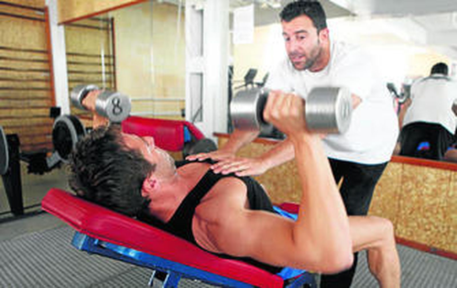 Un monitor ayuda a un usuario de un gimnasio a realizar un ejercicio de pecho con unas mancuernas.