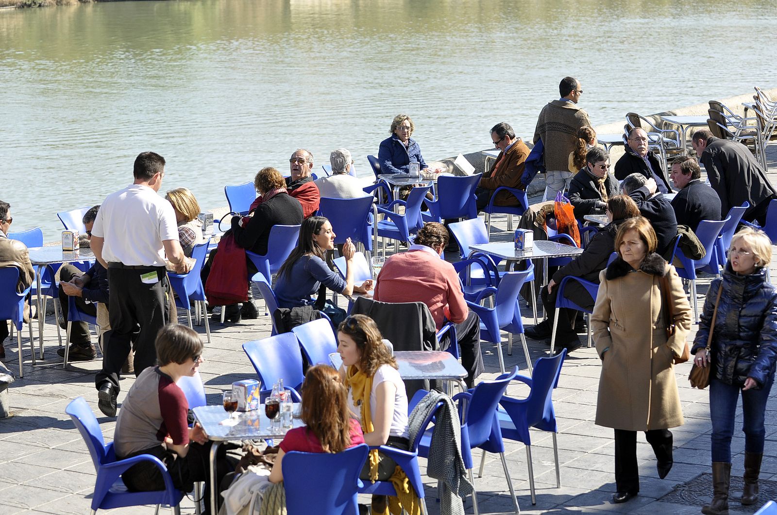 Una terraza de un bar junto al río abarrotada en un día soleado.