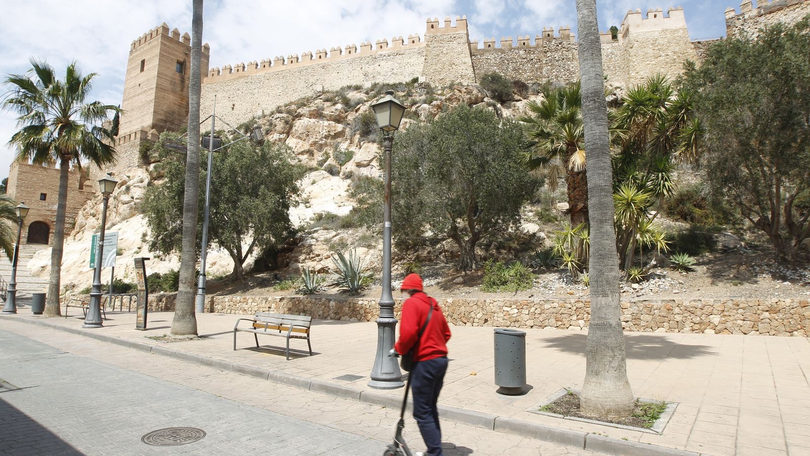 Un joven, con mascarilla y guantes, circula en patinete por delante de la Alcazaba
