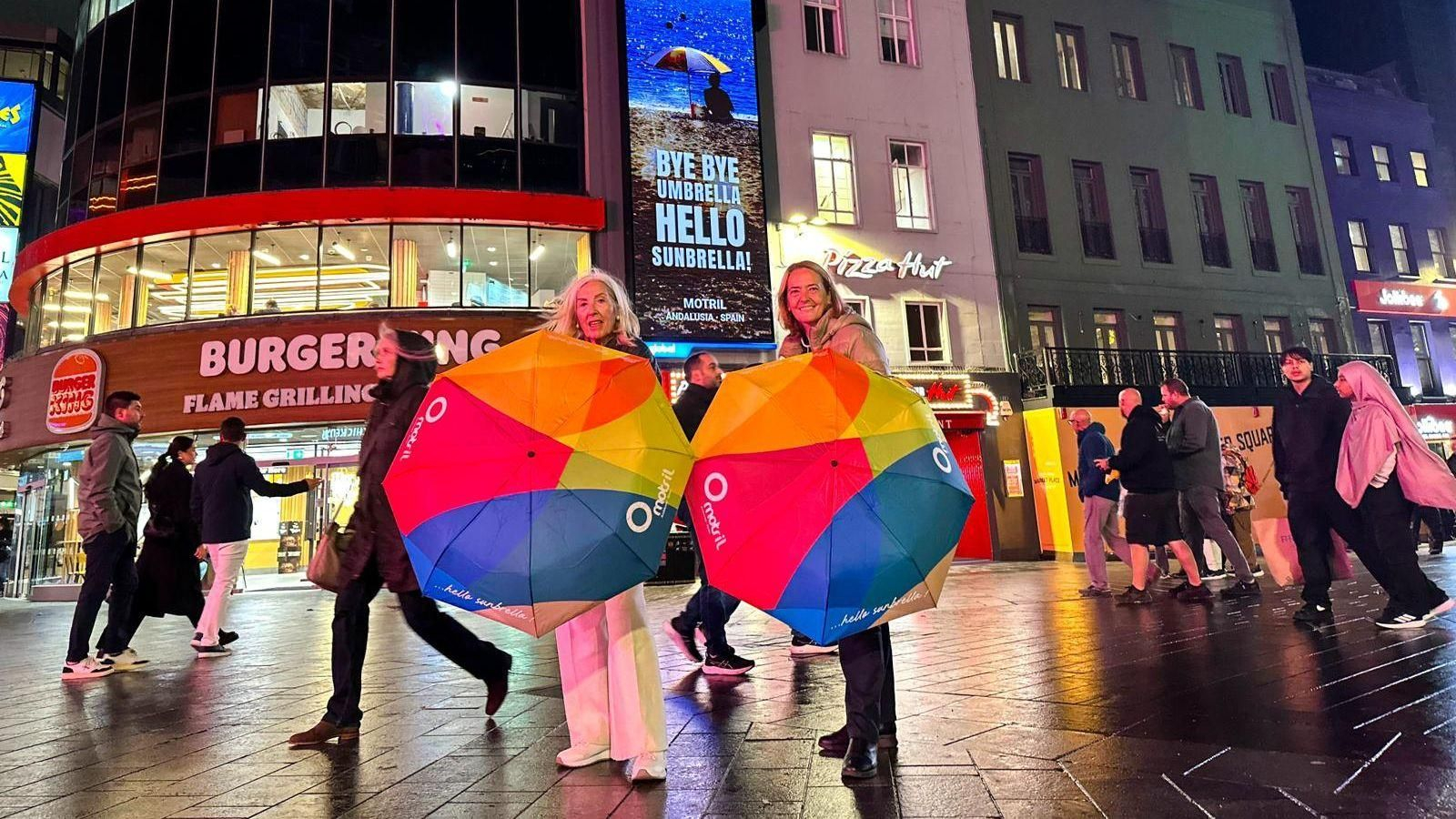 Luisa García y María Ángeles Escámez frente a la campaña “Adiós sombrilla, hola parasol” en las pantallas de la calle Leicester Square