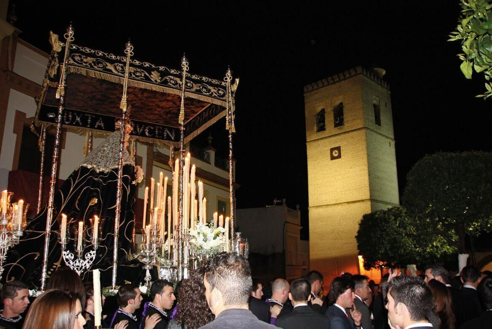La Virgen de los Dolores en la Plaza del Calvario bajo su majestuoso palio de la Bajada con piezas del siglo XVIII y XIX. Al fondo, la torre de la parroquia.