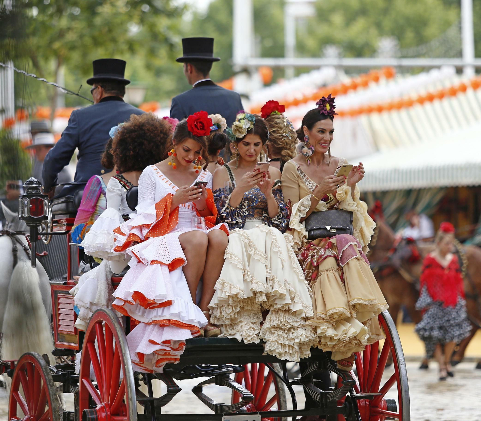 Mujeres de flamenca consultan el móvil mientras pasean en coche de caballo.