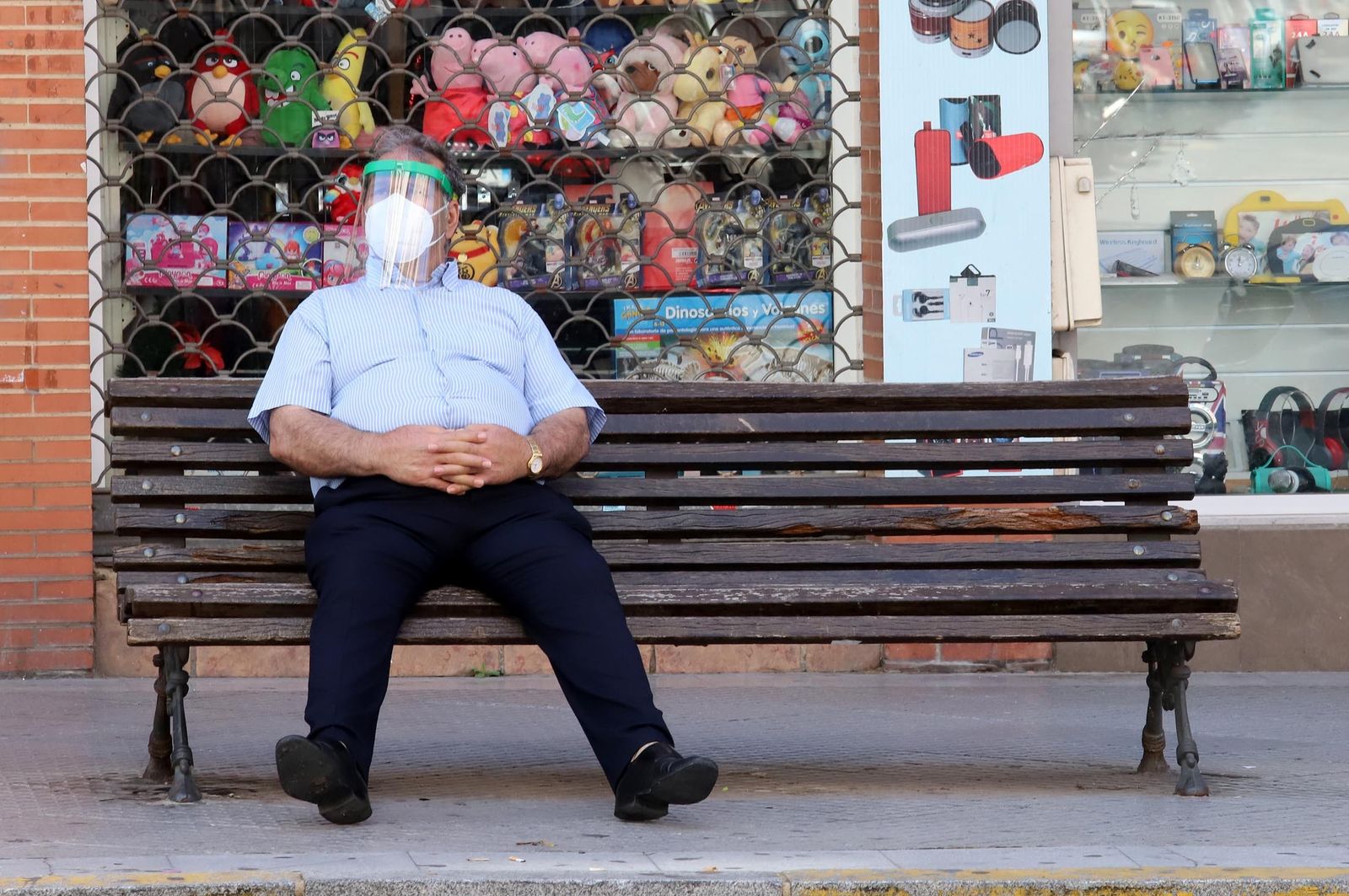 Un hombre se sienta en un banco con una mascarilla y una pantalla de protección.