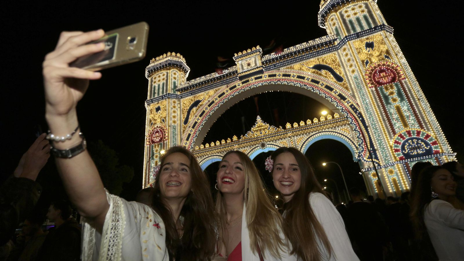 Tres jóvenes se fotografían delante de la portada de la Feria de Abril de 2018 el día del alumbrado.