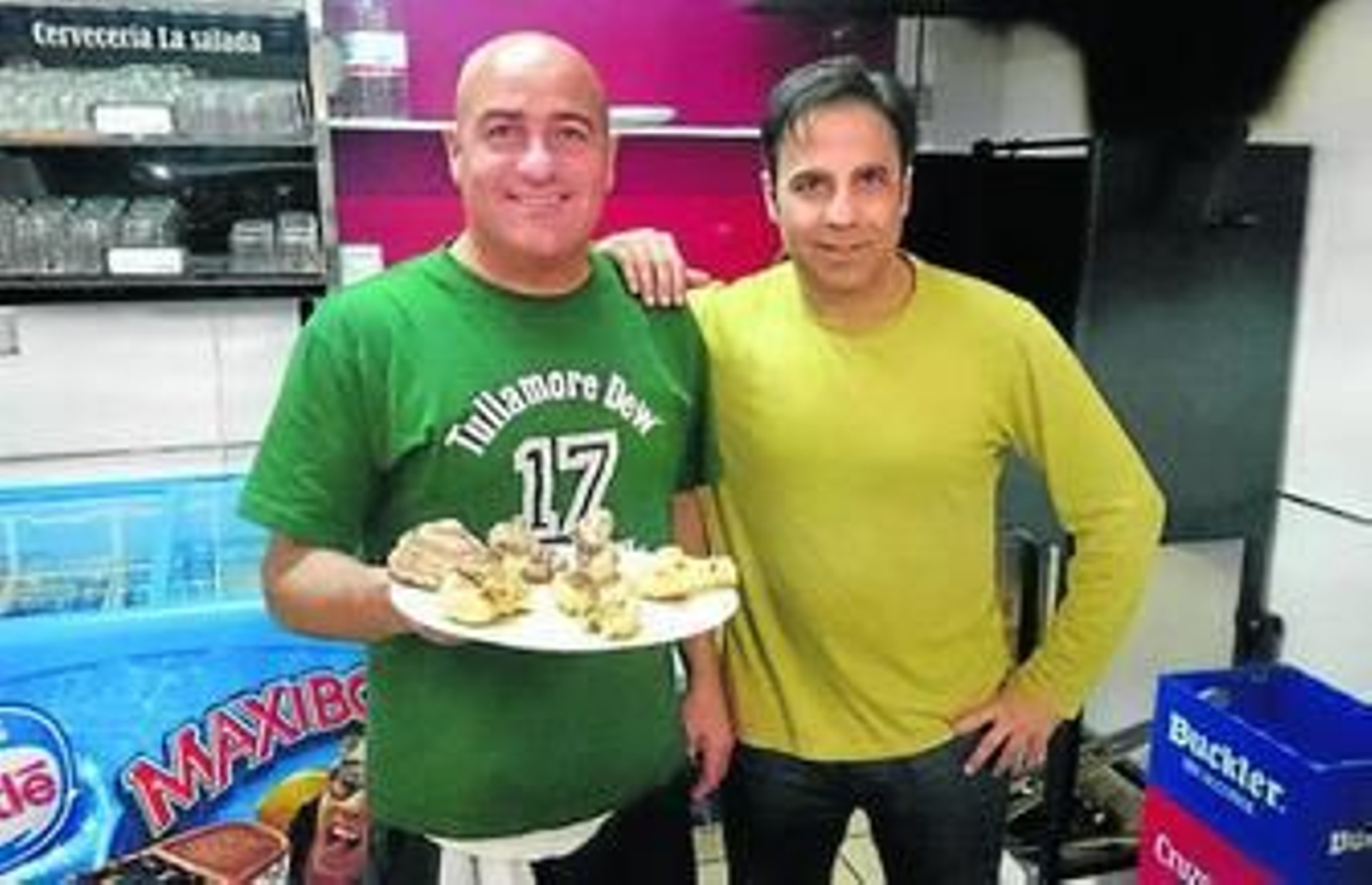 Juan y Bernardo posan detrás de la barra del ambigú con unas tapas de costilla y alitas de pollo.